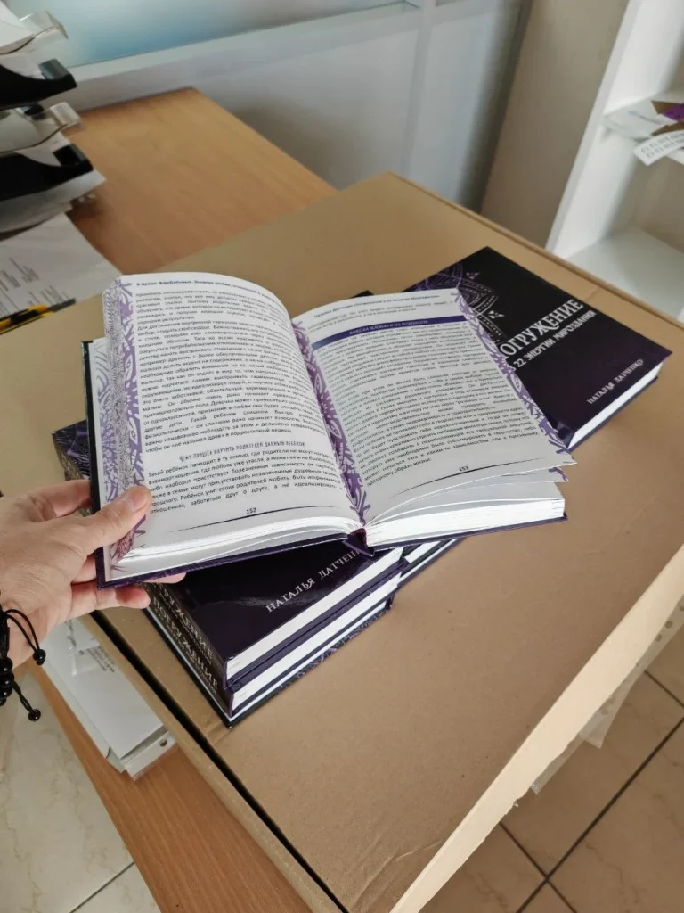 Розгорнута книжка з фіолетовим оформленням сторінок на тлі тиражу книжок у твердій палітурці
